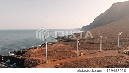 Tenerife: White wind turbines working on dry, rocky coastline, producing wind power renewable and sustainable energy, with the serene ocean and sunset mountains in background. Aerial drone flight Tenerife: White wind turbines working on dry, rocky coastline, producing wind power renewable and sustainable energy, with the serene ocean and sunset mountains in background. Aerial drone flight 139488938