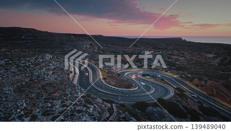 Malta: Aerial view of cars driving on a winding road on the coast of Malta at bright sunset, creating a scenic landscape with the Mediterranean Sea in the background. Drone flight Malta: Aerial view of cars driving on a winding road on the coast of Malta at bright sunset, creating a scenic landscape with the Mediterranean Sea in the background. Drone flight 139489040