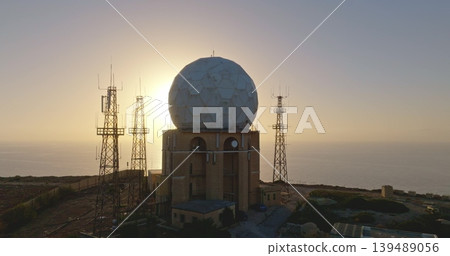Large radar antenna and communication towers dominate the landscape of a coastal military installation, silhouetted against the warm hues of a setting sun over the calm sea. Drone flight Large radar antenna and communication towers dominate the landscape of a coastal military installation, silhouetted against the warm hues of a setting sun over the calm sea. Drone flight 139489056