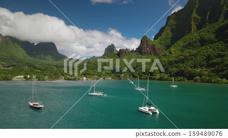 Sailing boats anchoring in a beautiful turquoise lagoon, surrounded by lush green tropical mountains and a small village on the coast of French Polynesia Sailing boats anchoring in a beautiful turquoise lagoon, surrounded by lush green tropical mountains and a small village on the coast of French Polynesia 139489076