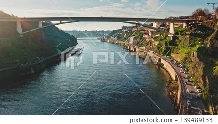 Portugal, Porto: Douro River with cruise boats, bridges and car traffic moving along the riverbank, showcasing urban city landscape with green hills under clear sky. Aerial view drone Portugal, Porto: Douro River with cruise boats, bridges and car traffic moving along the riverbank, showcasing urban city landscape with green hills under clear sky. Aerial view drone 139489133