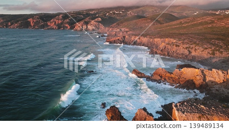 Portugal, Lisbon: Atlantic ocean coast waves crashing onto rugged cliffs of Sintra Cascais Natural Park in golden sunset hour light, aerial view landscape. Natural landscape, travel background Portugal, Lisbon: Atlantic ocean coast waves crashing onto rugged cliffs of Sintra Cascais Natural Park in golden sunset hour light, aerial view landscape. Natural landscape, travel background 139489134