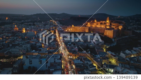 Malta, Comino island: Aerial view illuminated historic fortified Cittadella. Modern city buildings and streets glowing over Victoria on Gozo during enchanting twilight hours. Drone flight 139489139