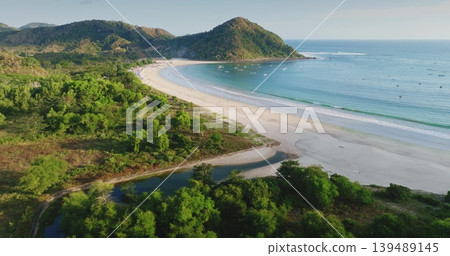 Lombok, Selong Belanak Beach presents its white sand, tranquil turquoise waters, and lush green hills, with traditional fishing boats dotting the bay. Tropical travel paradise background, aerial view 139489145