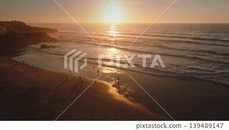 Portugal, Lisbon: People enjoy sunset on Praia do Guincho beach, walking along sandy shoreline, bright orange sun reflects on the Atlantic Ocean waves. Summer relaxation. Aerial drone flight Portugal, Lisbon: People enjoy sunset on Praia do Guincho beach, walking along sandy shoreline, bright orange sun reflects on the Atlantic Ocean waves. Summer relaxation. Aerial drone flight 139489147