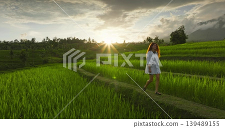Woman enjoying peaceful sunset stroll on pathways of lush green rice terraces on Bali island, experiencing nature and serenity during a travel adventure in Indonesia. Beautiful tropical vacation Woman enjoying peaceful sunset stroll on pathways of lush green rice terraces on Bali island, experiencing nature and serenity during a travel adventure in Indonesia. Beautiful tropical vacation 139489155