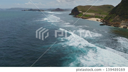 Blue ocean waves rolling towards the sandy beach and rocky coastline of Bukit Merese in Lombok, Indonesia, green hills rising in the background under cloudy sky. Nature landscape. Aerial drone flight Blue ocean waves rolling towards the sandy beach and rocky coastline of Bukit Merese in Lombok, Indonesia, green hills rising in the background under cloudy sky. Nature landscape. Aerial drone flight 139489169