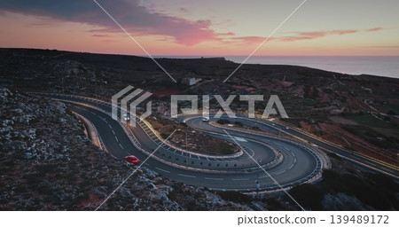 Malta: Winding road with cars driving along the coast of Malta during a beautiful sunset with pink and purple clouds, creating a scenic travel destination view. Aerial drone flight 139489172
