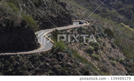 Tenerife, Canary Islands: Aerial view of car navigating winding mountain road with safety barriers, highlighting rocky dramatic volcanic landscape. Drone flight 139489179