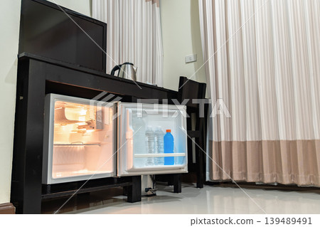 A small refrigerator sits beneath a wooden desk with chair, creating a clean hotel room setup that supports minibar service and compact guest comfort. 139489491