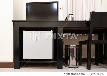 A small refrigerator sits beneath a wooden desk with chair, creating a clean hotel room setup that supports minibar service and compact guest comfort. A small refrigerator sits beneath a wooden desk with chair, creating a clean hotel room setup that supports minibar service and compact guest comfort. 139489492