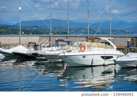 Modern motorboats lined up in marina on Lake Garda, Italy, reflecting in water, with distant mountains and summer sky creating leisure travel mood Modern motorboats lined up in marina on Lake Garda, Italy, reflecting in water, with distant mountains and summer sky creating leisure travel mood 139490091