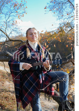 Blonde woman playing guitar ukulele outdoors in autumn forest. Concept of sound therapy, mental health and wellness rituals. Calmness tranquility audio-sensory practices. Aura farming Blonde woman playing guitar ukulele outdoors in autumn forest. Concept of sound therapy, mental health and wellness rituals. Calmness tranquility audio-sensory practices. Aura farming 139490764