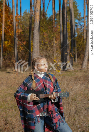 Portrait of smiling caucasian girl plays ukulele in autumnal park. Young woman plays guitar musical instrument outside in nature fall time. Audio music healing Portrait of smiling caucasian girl plays ukulele in autumnal park. Young woman plays guitar musical instrument outside in nature fall time. Audio music healing 139490765