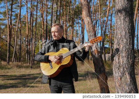 Blonde man playing string guitar outdoors in autumn forest. Concept of sound therapy, mental health and wellness rituals. Calmness tranquility audio-sensory practices. Aura farming energy Blonde man playing string guitar outdoors in autumn forest. Concept of sound therapy, mental health and wellness rituals. Calmness tranquility audio-sensory practices. Aura farming energy 139490853