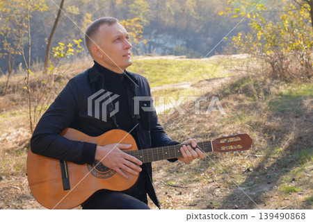 Beautiful blonde man playing acoustic guitar and singing relaxing enjoying outdoors on sunny autumn day. Artist learning and practicing playing musical instrument. Fall leaves autumnal Beautiful blonde man playing acoustic guitar and singing relaxing enjoying outdoors on sunny autumn day. Artist learning and practicing playing musical instrument. Fall leaves autumnal 139490868