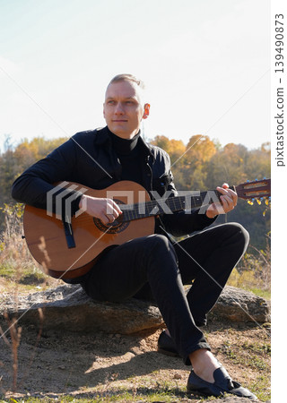 Portrait of smiling caucasian artist plays wooden acoustic guitar in autumnal park. Young songwriter plays string musical instrument hobby outside in nature fall time. Audio music healing Portrait of smiling caucasian artist plays wooden acoustic guitar in autumnal park. Young songwriter plays string musical instrument hobby outside in nature fall time. Audio music healing 139490873