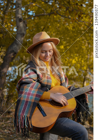 Beautiful blonde girl playing acoustic guitar and singing relaxing enjoying outdoors on sunny autumn day. Hippie woman in boho style learning and practicing playing musical instrument. Fall leaves Beautiful blonde girl playing acoustic guitar and singing relaxing enjoying outdoors on sunny autumn day. Hippie woman in boho style learning and practicing playing musical instrument. Fall leaves 139490877