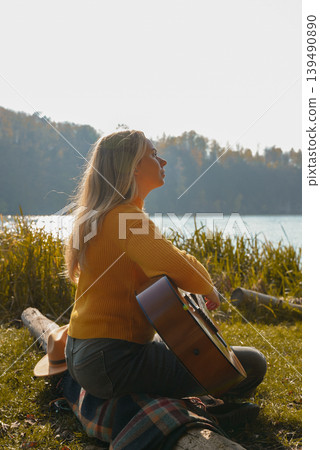 Beautiful blonde girl playing acoustic guitar and singing relaxing enjoying outdoors on sunny autumn day. Hippie woman in boho style learning and practicing playing musical instrument. Fall leaves Beautiful blonde girl playing acoustic guitar and singing relaxing enjoying outdoors on sunny autumn day. Hippie woman in boho style learning and practicing playing musical instrument. Fall leaves 139490890