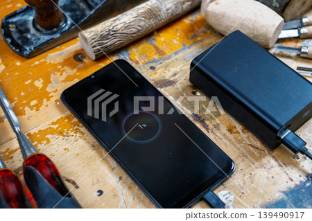 Close-up of a phone screen showing 1% charge, connected to a power bank among wood carving tools and blanks. Close-up of a phone screen showing 1% charge, connected to a power bank among wood carving tools and blanks. 139490917