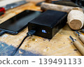 Close-up of a black power bank showing a 100% digital charge indicator, connected to a smartphone on a rustic wooden workshop table. 139491133