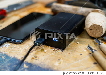 Close-up of a black power bank showing a 100% digital charge indicator, connected to a smartphone on a rustic wooden workshop table. 139491133