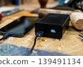 Close-up of a black power bank showing a 100% digital charge indicator, connected to a smartphone on a rustic wooden workshop table. 139491134