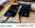 Close-up of a black power bank showing a 100% digital charge indicator, connected to a smartphone on a rustic wooden workshop table. 139491135