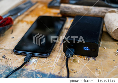 Close-up of a black power bank showing a 100% digital charge indicator, connected to a smartphone on a rustic wooden workshop table. Close-up of a black power bank showing a 100% digital charge indicator, connected to a smartphone on a rustic wooden workshop table. 139491135