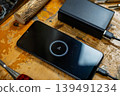 Close-up of a phone screen showing 2% charge, connected to a power bank among wood carving tools and logs. 139491234