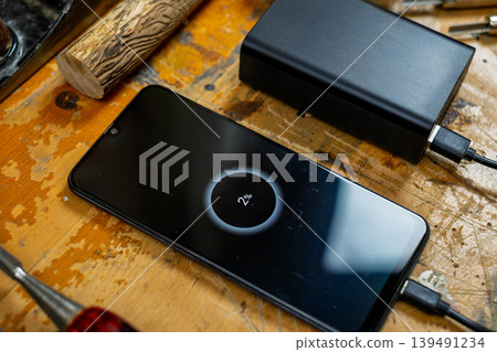 Close-up of a phone screen showing 2% charge, connected to a power bank among wood carving tools and logs. Close-up of a phone screen showing 2% charge, connected to a power bank among wood carving tools and logs. 139491234