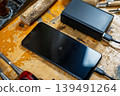Close-up of a phone screen showing 3% charge, connected to a power bank among wood carving tools and timber. 139491264