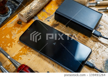 Close-up of a phone screen showing 3% charge, connected to a power bank among wood carving tools and timber. Close-up of a phone screen showing 3% charge, connected to a power bank among wood carving tools and timber. 139491264