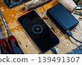 Angled shot of a smartphone screen showing 3% battery, connected to a power bank among chisels and wood blanks on a carpenter's table. 139491307