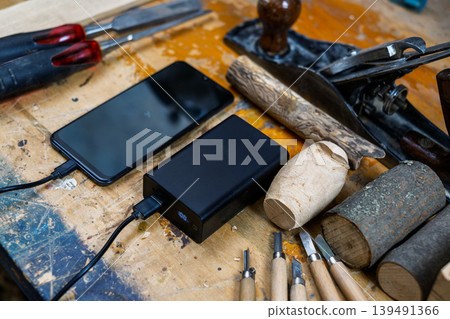 A wide high-angle shot of a phone connected to a 100% charged power bank, surrounded by chisels, wood planes, and logs. 139491366