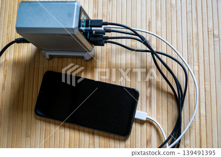 A high-tech silver charging hub with multiple cables connected to a smartphone on a natural bamboo surface. 139491395