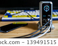 A silver charging hub displaying 11W output, connected to a smartphone and other devices on a desk with a mechanical keyboard. 139491515