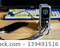 A silver charging hub displaying 11W output, connected to a smartphone and other devices on a desk with a mechanical keyboard. 139491516