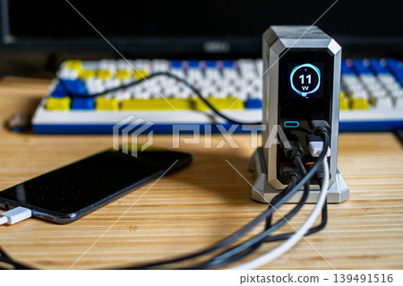 A silver charging hub displaying 11W output, connected to a smartphone and other devices on a desk with a mechanical keyboard. A silver charging hub displaying 11W output, connected to a smartphone and other devices on a desk with a mechanical keyboard. 139491516