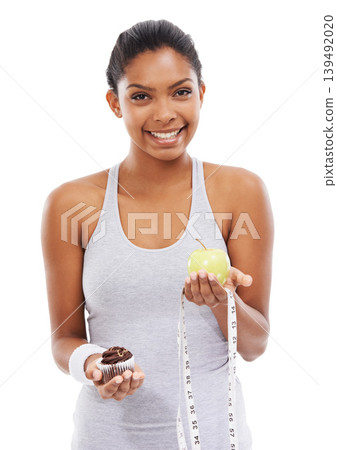 Portrait, woman and choice of apple or cupcake in studio isolated on a white background to lose weight. Smile, fitness or exercise with a happy young athlete holding fruit, dessert and measuring tape Portrait, woman and choice of apple or cupcake in studio isolated on a white background to lose weight. Smile, fitness or exercise with a happy young athlete holding fruit, dessert and measuring tape 139492020
