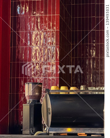 A modern coffee machine sits on a counter against a glossy red tiled wall. The machine is sleek and black, with yellow cups stacked on top. 139493831