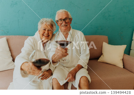 Happy senior couple sitting together in bathrobe on sofa with glass of wine. Happy senior couple sitting together in bathrobe on sofa with glass of wine. 139494428