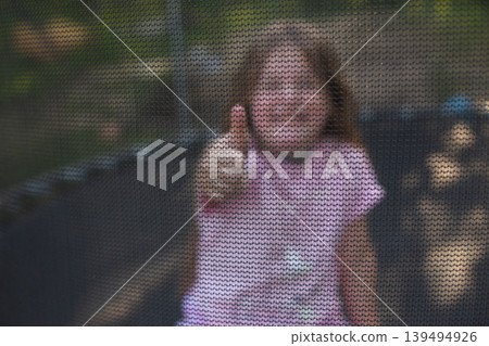 Happy excited child girl sitting on trampoline outdoor showing thumb up sign Happy excited child girl sitting on trampoline outdoor showing thumb up sign 139494926
