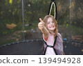 Happy excited child girl sitting on trampoline outdoor showing thumb up sign 139494928