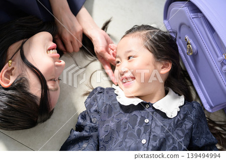 A Japanese parent and child are lying on the floor, smiling and gazing at each other with their school bags in hand. 139494994