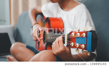 Asian young boy practicing acoustic guitar on sofa at home. Child learning musical instrument, hobby and lifestyle concept for education and talent development. Asian young boy practicing acoustic guitar on sofa at home. Child learning musical instrument, hobby and lifestyle concept for education and talent development. 139495143