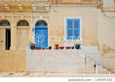 Sunny facade of historic Malta limestone building with bright blue wooden door, matching shutters, ornate ironwork arch, terracotta and blue flower pots on white wall Sunny facade of historic Malta limestone building with bright blue wooden door, matching shutters, ornate ironwork arch, terracotta and blue flower pots on white wall 139495192