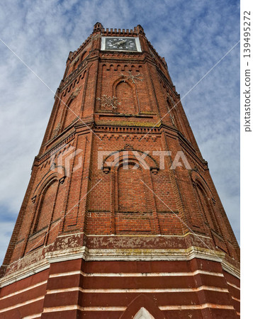 A tall brick water tower features a clock at the top. The sky is blue with white clouds. The shot captures the tower's height and detailed brickwork from below. 139495272