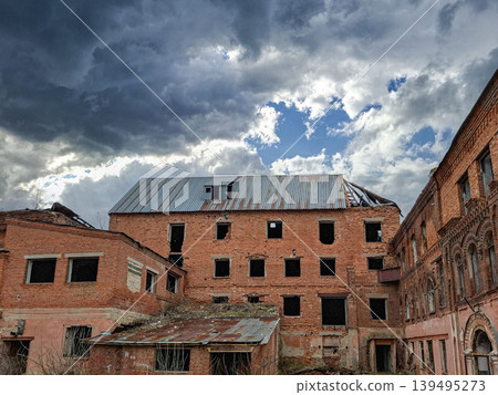 A red brick tobacco factory shows signs of decay with broken windows and an open roof. The sky is filled with dark clouds, creating a stark contrast to the building's structure. A red brick tobacco factory shows signs of decay with broken windows and an open roof. The sky is filled with dark clouds, creating a stark contrast to the building's structure. 139495273