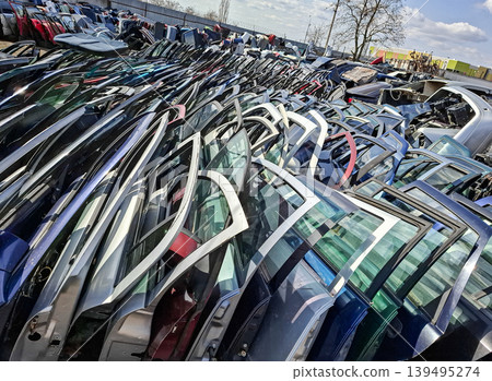 At a vehicle recycling center, a large pile of used car doors sits under the bright sun. The doors are from various cars and show signs of wear. This scene captures the recycling process. 139495274